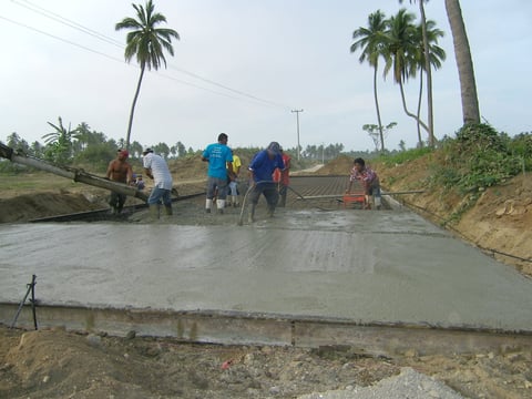 Workers smoothing fresh concrete on a construction site with palm trees and tropical landscape in background
