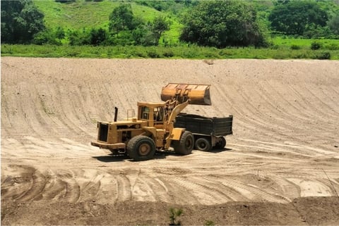 Yellow construction machinery with water tank working on dirt field with green hillside in background