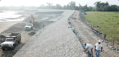 Sitio de construcción en ribera de río con trabajadores, camiones y terraplén de grava