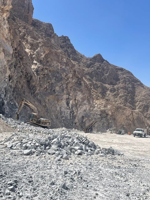 Operación de cantera al pie de montaña rocosa con excavadora y camión de volteo entre pilas de piedra extraída