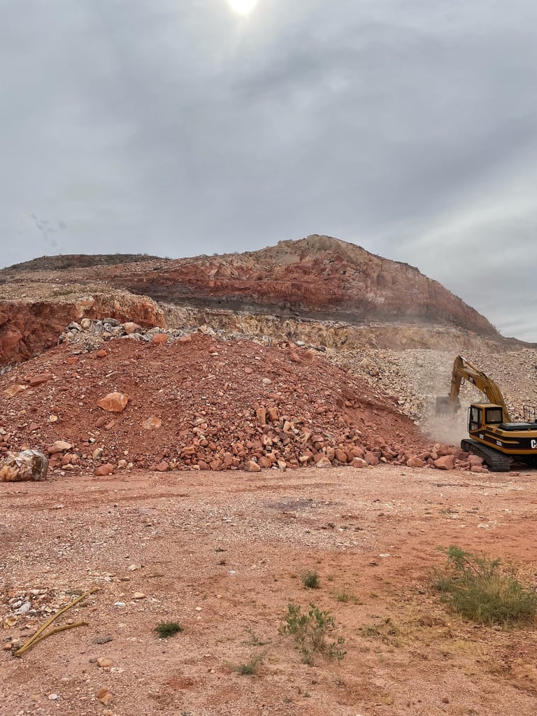 Excavator operating at a dusty mining or quarry site with large rocky earth mounds under cloudy sky