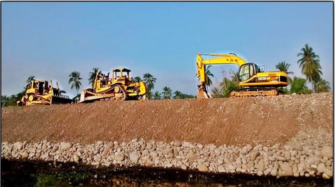 Heavy construction equipment including a yellow excavator and bulldozer working on an earthen embankment with palm trees in the background